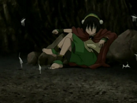 A young woman is sat in a cave wearing a green outfit and a red cape. She is surrounded by half a dozen small, white insects resembling grasshoppers jumping in the air.