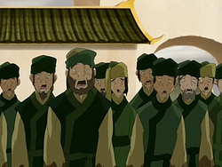 A large group of people in green outfits are walking forward, their mouths a gape and with a dazed expression on their faces. Behind them is a building with a green angled roof.